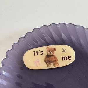 Détail d'une pince à cheveux ours en résine avec texte It's Me dans un plateau violet