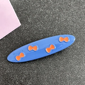 Detail shot of a blue oval hair clip featuring orange bow prints and polished metal finish