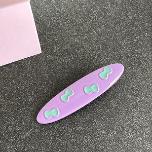 Close-up of lavender oval hair clip with mint green bow pattern on grey textured background