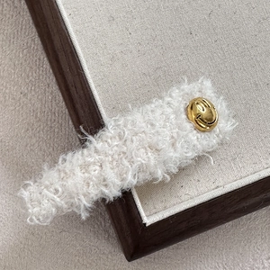 Detailaufnahme der weißen Plüsch-Haarklammer mit goldenem Smiley-Metallemblem-Detail