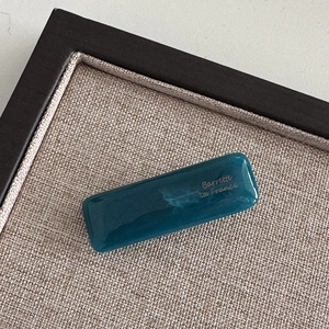 Detailed view of glossy dark teal French barrette with gold lettering on grey texture