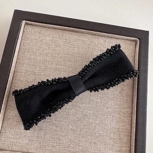 Elegant black bow hair clip with beaded edges and premium woven fabric on beige linen base