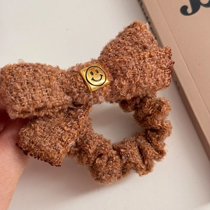 Detail view of a brown fleece bow hair tie featuring a golden square smiley face charm