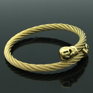 Detalle de brazalete rígido de cable de acero dorado con dos dijes en forma de calavera