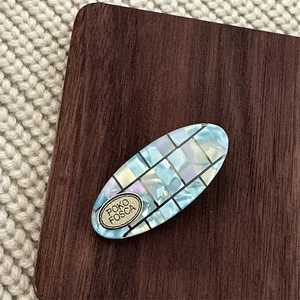 Detailed close-up of oval hair clip with iridescent blue mosaic pattern and gold logo