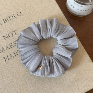 Close-up of a grey pleated silk scrunchie with a gold metal tag on a textured background