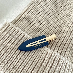 Detailed view of deep blue hair clip with gold logo on beige knit fabric background