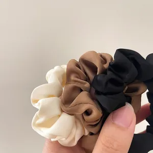 Close-up of three satin scrunchies in cream, brown, and black held in hand with soft silk texture