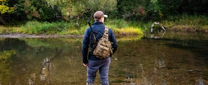 Khaki outdoor fishing tackle sling bag with Molle system shown in a scenic stream setting