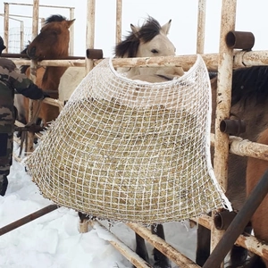 White nylon slow feeder hay bag with mesh design hanging on a stable fence in snow