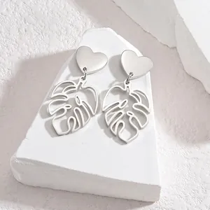 Close-up of silver stainless steel earrings with heart studs and monstera leaf pendants