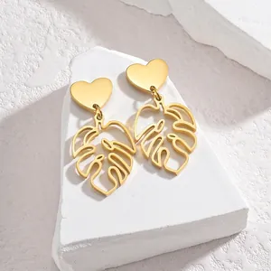 Detailed close-up of golden heart studs with hollow monstera leaf pendant on white stone