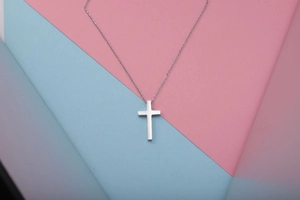 Gepolijste zilveren kruishanger aan fijne O-ketting op roze en blauwe achtergrond
