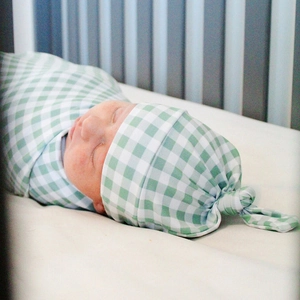 Detail view of a green gingham baby swaddle blanket and matching knotted hat in a crib