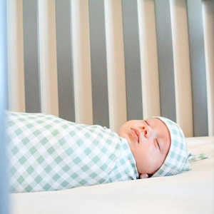 Close-up of green gingham baby swaddle blanket and matching hat on a wooden crib