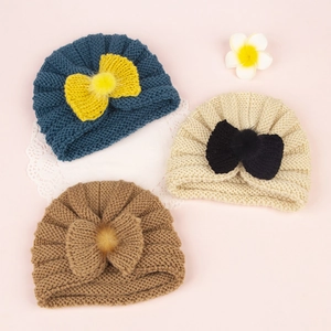 Three wave-knit baby turban hats with large bows and soft pom-poms on a pink background