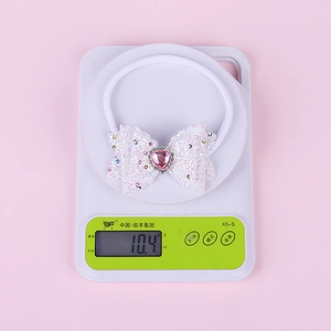 Detail view of a white sequin bow headband with a pink heart gem on an electronic scale