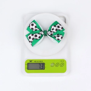 Macro view of a soccer-themed bow hair clip on a scale showing a light weight of 7.3 grams