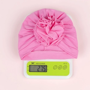 Pink cotton baby turban hat with flower detail on scale showing 27.5g weight