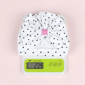 Vue détaillée du bonnet en coton pour bébé de 23,4g avec nœud à pois roses sur une balance