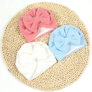 Three soft dotted velvet baby hats with cute bow details on a natural woven mat background