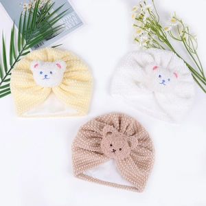 Close-up of three waffle knit baby hats with cute plush bear patches in neutral tones