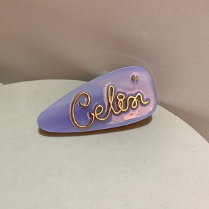 Dettaglio macro della molletta per capelli lilla in acetato con scritta oro e strass rosa