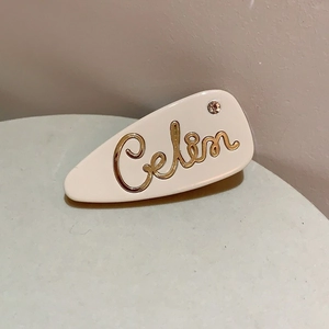 Dettaglio del fermaglio in acetato crema con scritta Celin dorata e strass brillante
