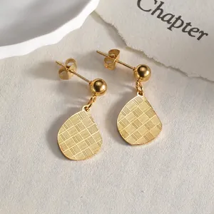 Close-up of golden teardrop earrings with textured grid patterns on a minimal paper backdrop