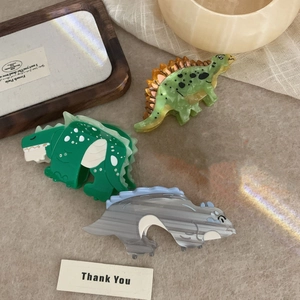 Three dinosaur-shaped acetate hair claws in green and grey on aesthetic warm fabric