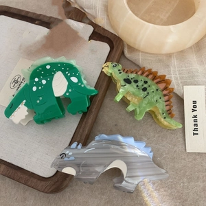 Three dinosaur-shaped acetate hair claws in green and grey textures on a wood tray