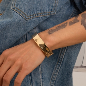 Dettaglio di un ampio bracciale a polsino in oro lucido indossato sulla mano e denim blu