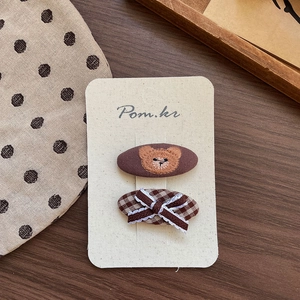 Detail view of brown fabric hair clips with teddy bear embroidery and a lace bow on a card