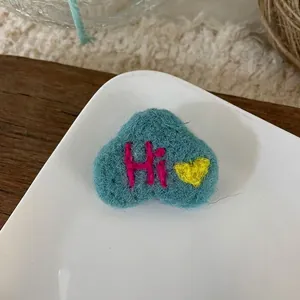 Detail of a handmade heart-shaped wool felt brooch in teal with embroidered Hi text