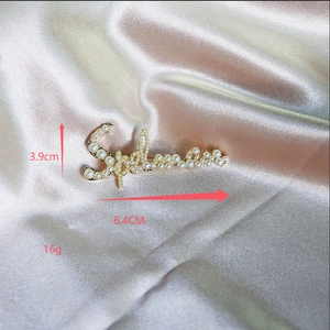 Dettaglio di una spilla dorata di lusso con scritta Supreme e perle bianche, lunga 8,4 cm