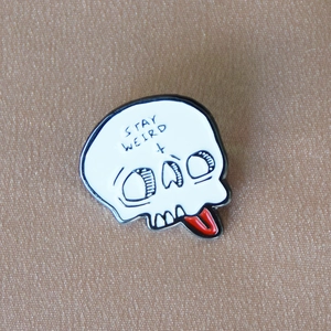 Detail of Stay Weird enamel skull pin with a red tongue on a textured fabric background