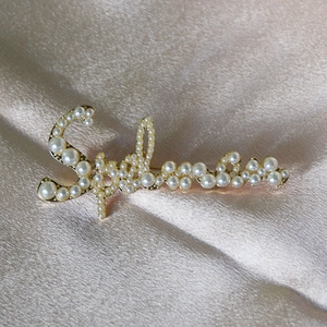 Elegante spilla a forma di lettera dorata con perle bianche su sfondo di raso champagne