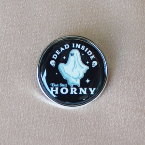 Detail of a round black enamel pin featuring a ghost and Dead Inside But Still Horny text