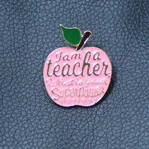 Detailed macro view of pink apple enamel teacher brooch with gold trim and superpower text