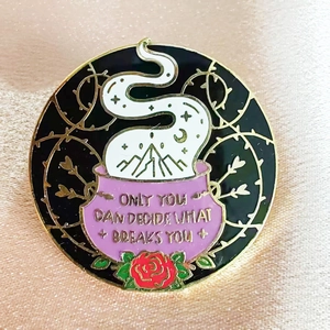 Macro detail of enamel pin with purple cauldron and inspirational quote on satin fabric
