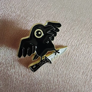 Close-up of a black crow holding a white knife enamel pin on a textured pink background