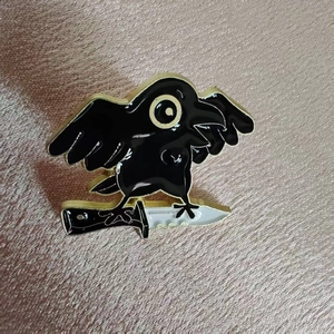 Cartoon raven standing on a dagger enamel pin with black finish and gold edges