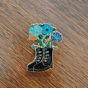 Dettaglio della spilla smaltata con stivaletti neri e fiori blu su fondo legno