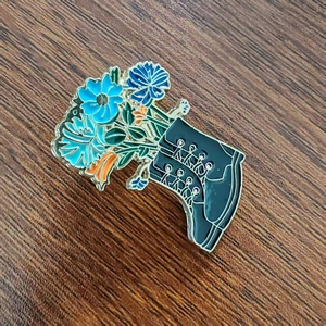 Dettaglio di una spilla creativa in smalto con stivali di pelle nera e bouquet di fiori blu