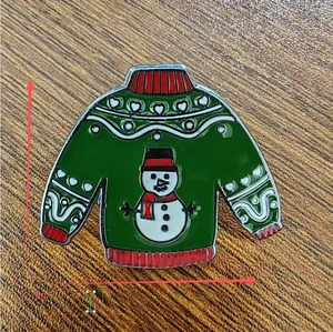 Detail of green snowman sweater enamel pin with dimensions 2.9cm by 3.5cm on wood