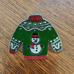 Close-up of a green Christmas sweater enamel pin with a snowman motif on wooden background