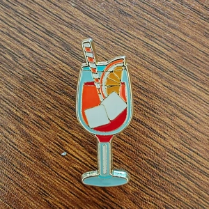 Detailed close-up of a cocktail enamel pin with red liquid and orange slice on wood