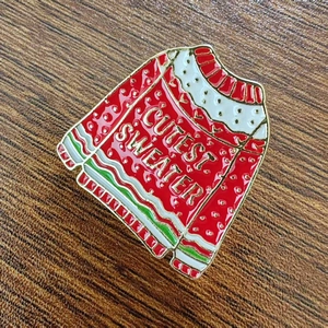 Detail of red enamel Christmas ugly sweater brooch with Cutest Sweater gold lettering