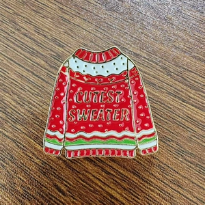 Festive red Christmas sweater enamel pin badge with Cutest Sweater text on wood background