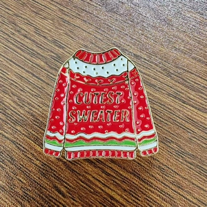 Macro detail of a red Christmas sweater enamel brooch with Cutest Sweater lettering